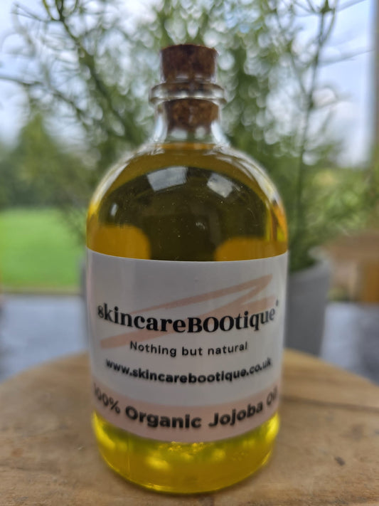 A glass bottle containing organic jojoba oil, with a label that includes the brand name 'skincarebootique' and the product name 'Jojoba Oil'. The bottle is placed in front of a plant, on a wooden surface.